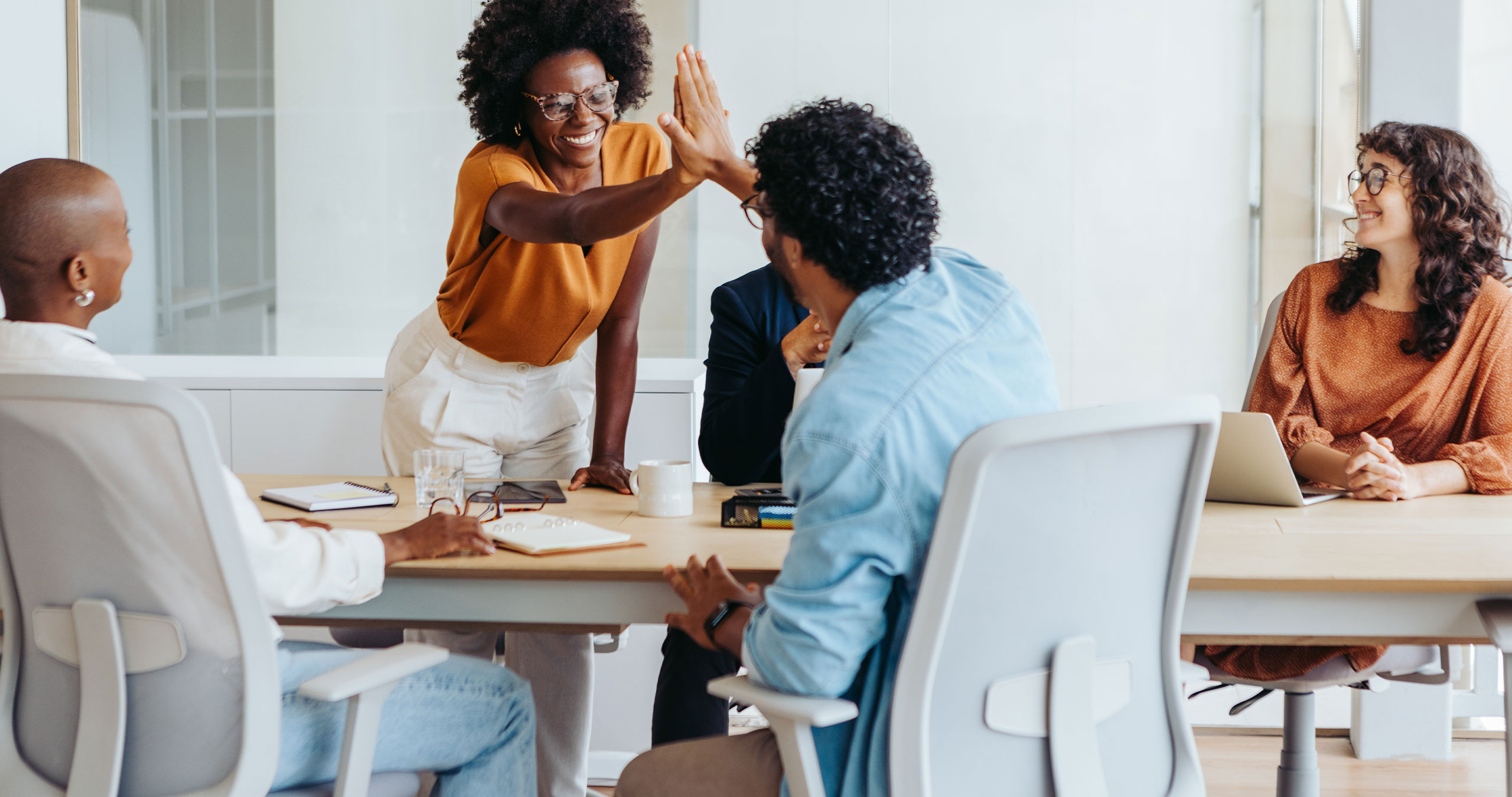 Five employees sit around a table, while two high-five one another.