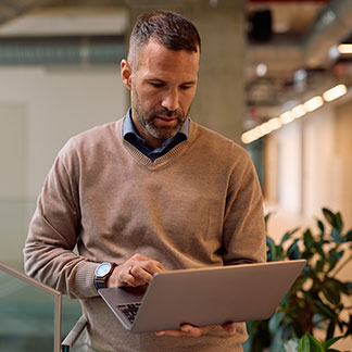 A man in a tan sweater holds his laptop in the office while taking notes.