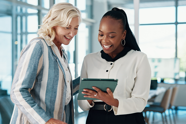Two women stand beside each other, talking as they look at a tablet at work.
